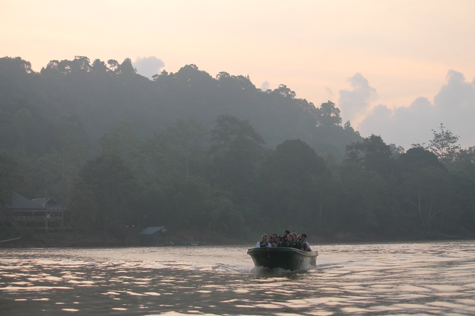 Kleines Boot mit Reisenden fährt bei Sonnenaufgang auf dem Kinabatangan River vor bewaldeten Hügeln in Sabah.