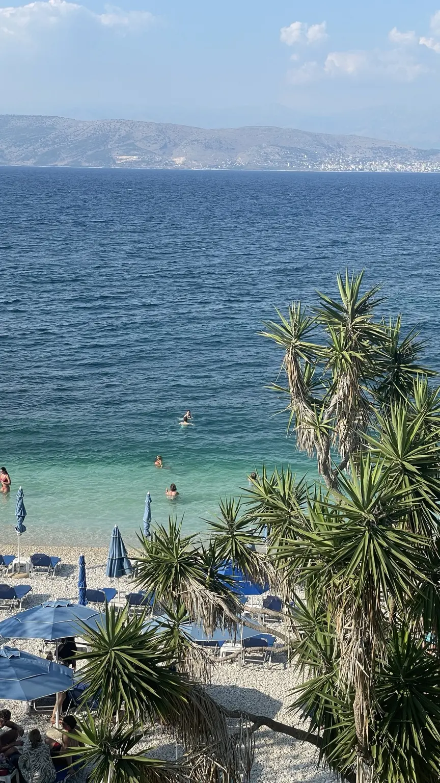 Kiesstrand bei Kassiopi mit blauen Sonnenschirmen und türkisfarbenem Wasser