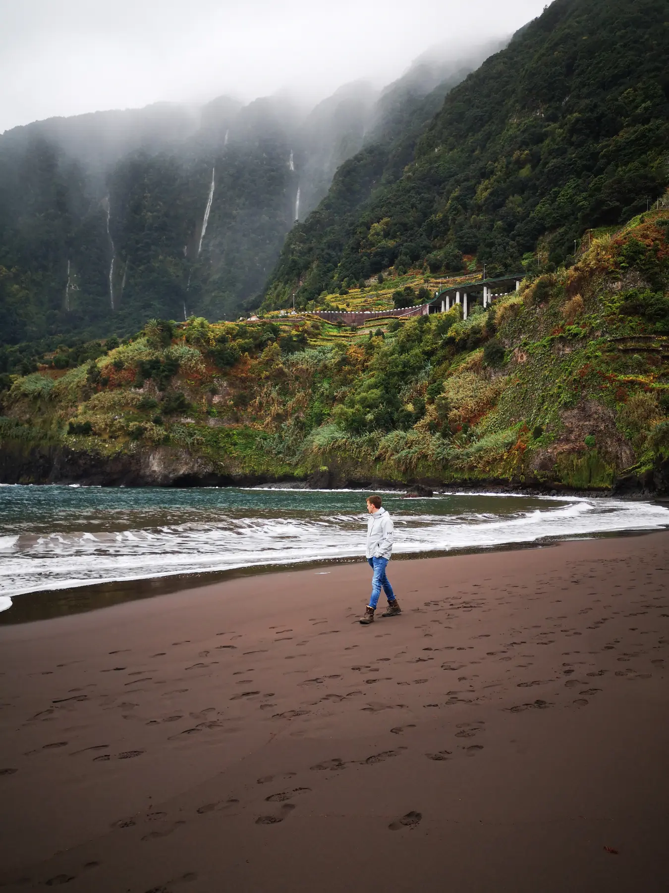 Wanderer am dunklen Sandstrand von Seixal vor grünen Berghängen und mehreren Wasserfällen auf Madeira