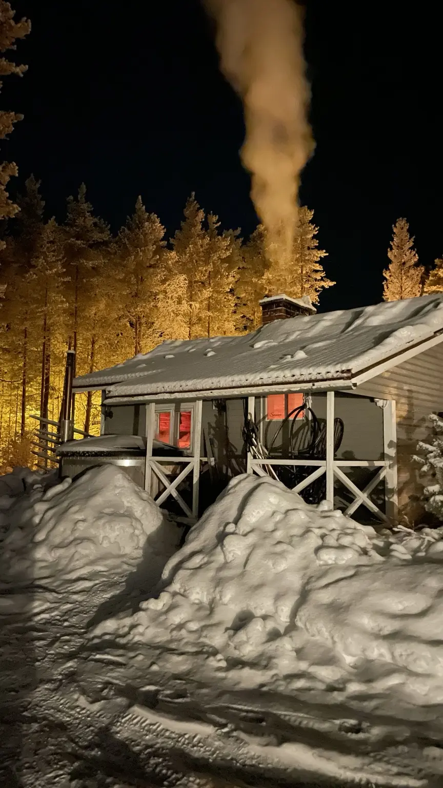 Tief verschneite Unterkunft im nachts beleuchteten Winterwald in Finnisch-Lappland