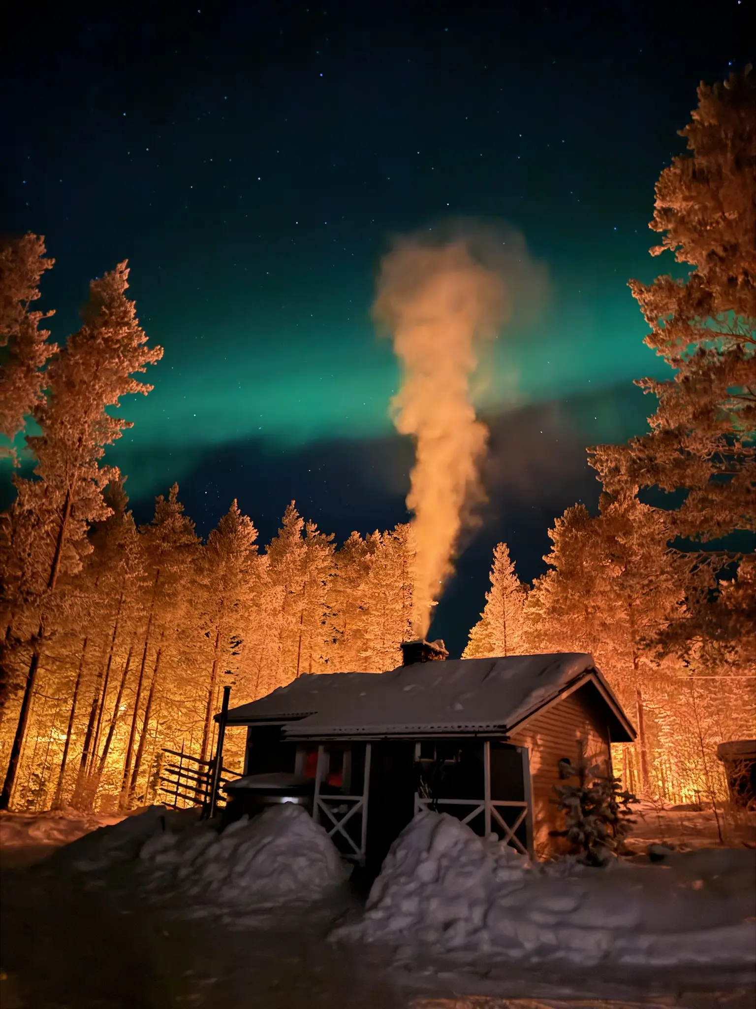Grüne Nordlichter über dem separaten Saunahaus im tief verschneiten Wald