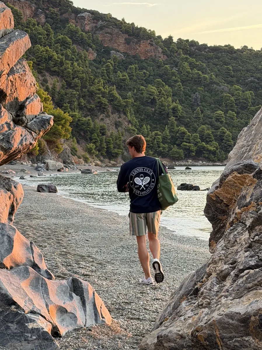 Person trägt den dunkelblauen Tennis-Club-Pullover an einem Strand auf Skopelos